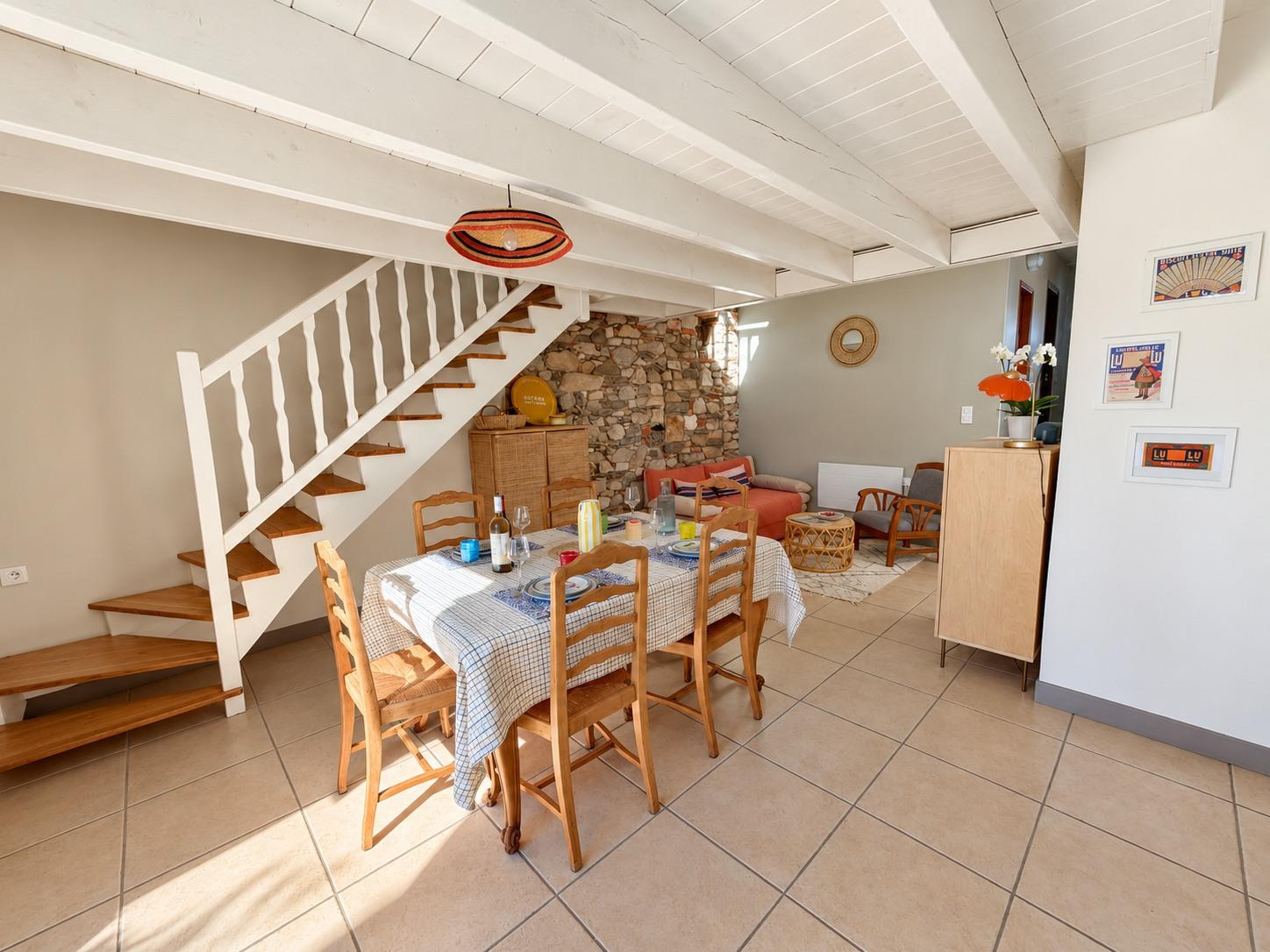 Le séjour de la maison principale : table en bois, escalier blanc et mur de pierre au fond